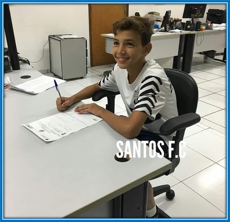 A Dream Realized: A beaming young Deivid captures the moment of triumph, seated with pen in hand, as he signs his first-ever contract with Santos, marking the inception of his professional football journey.